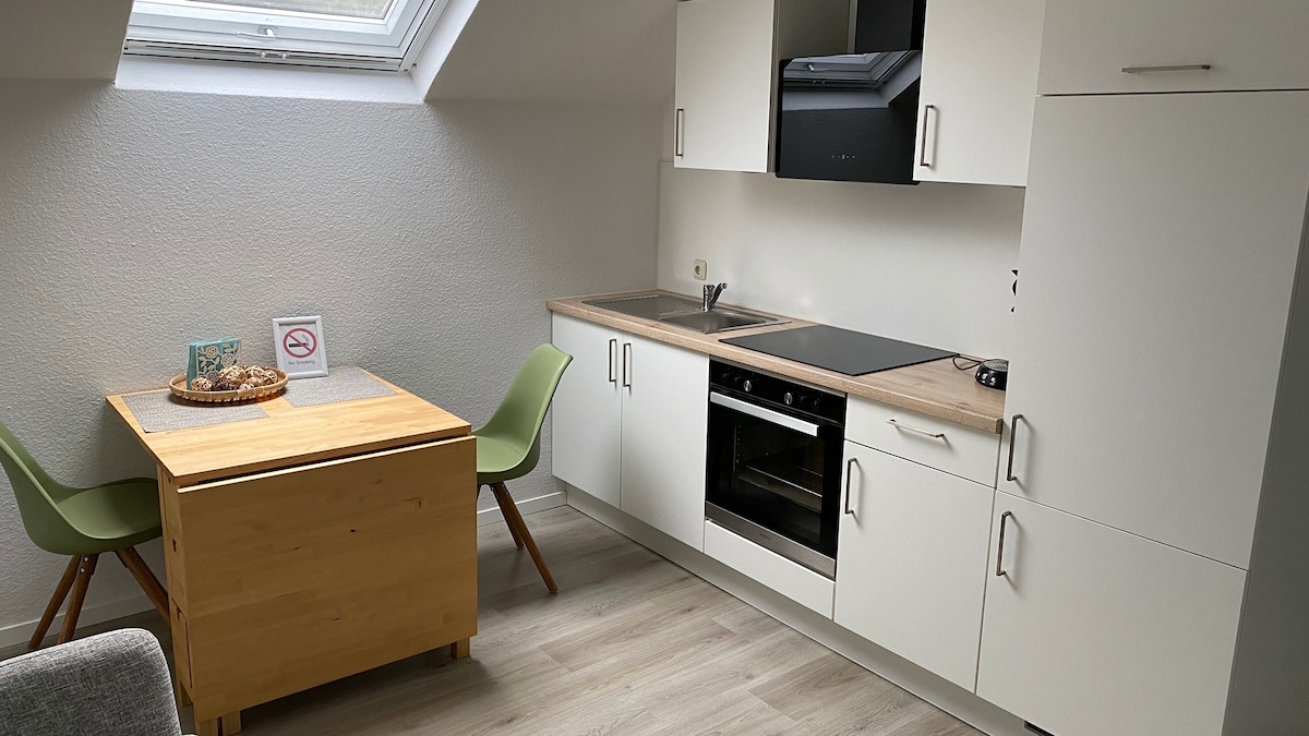 An open kitchen area features sleek white cabinetry and a modern oven. A wooden dining table is paired with two green chairs. Natural light enters through a skylight, creating a bright and functional space.