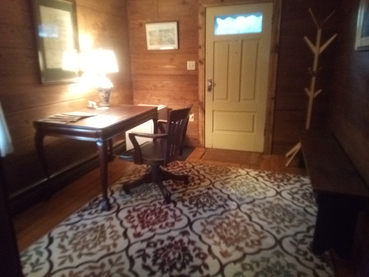 A warm corner of the room features a dark wood desk with a matching chair, accompanied by a table lamp that provides soft lighting. A colorful patterned rug covers the wooden floor, and a coat rack stands by the entrance door.