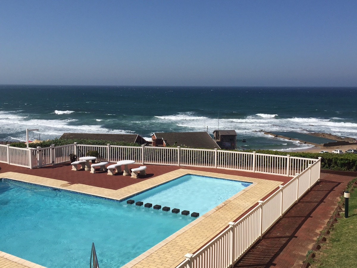 A clear view of the sparkling blue swimming pool is featured, bordered by a white railing. The ocean stretches in the background, revealing rolling waves under a bright sky. Sun loungers are positioned along the poolside, inviting relaxation.