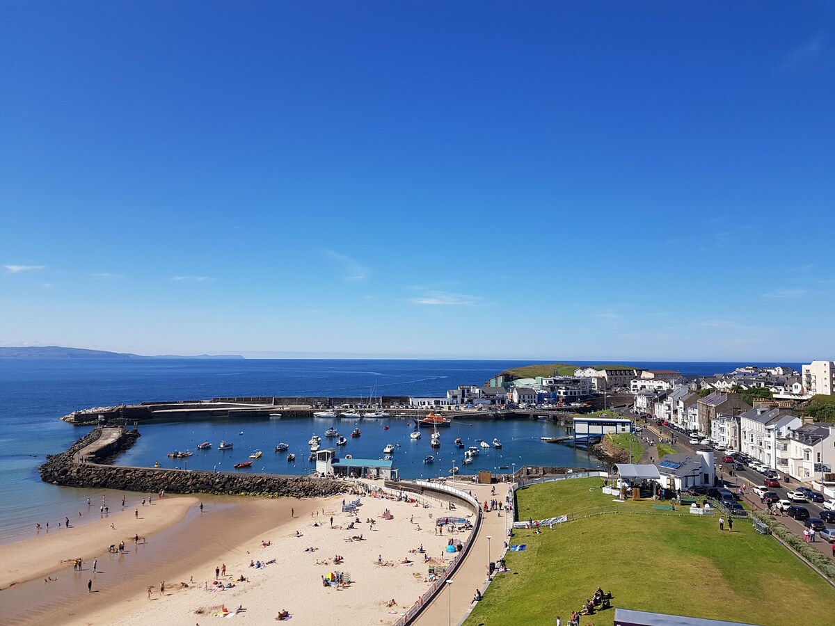 A coastal view displays a sandy beach leading to calm waters, dotted with small boats. The shoreline is occupied by sunbathers, while the adjacent harbor features waterfront buildings and lush greenery. A clear blue sky complements the serene atmosphere, highlighting the beauty of the seaside setting.