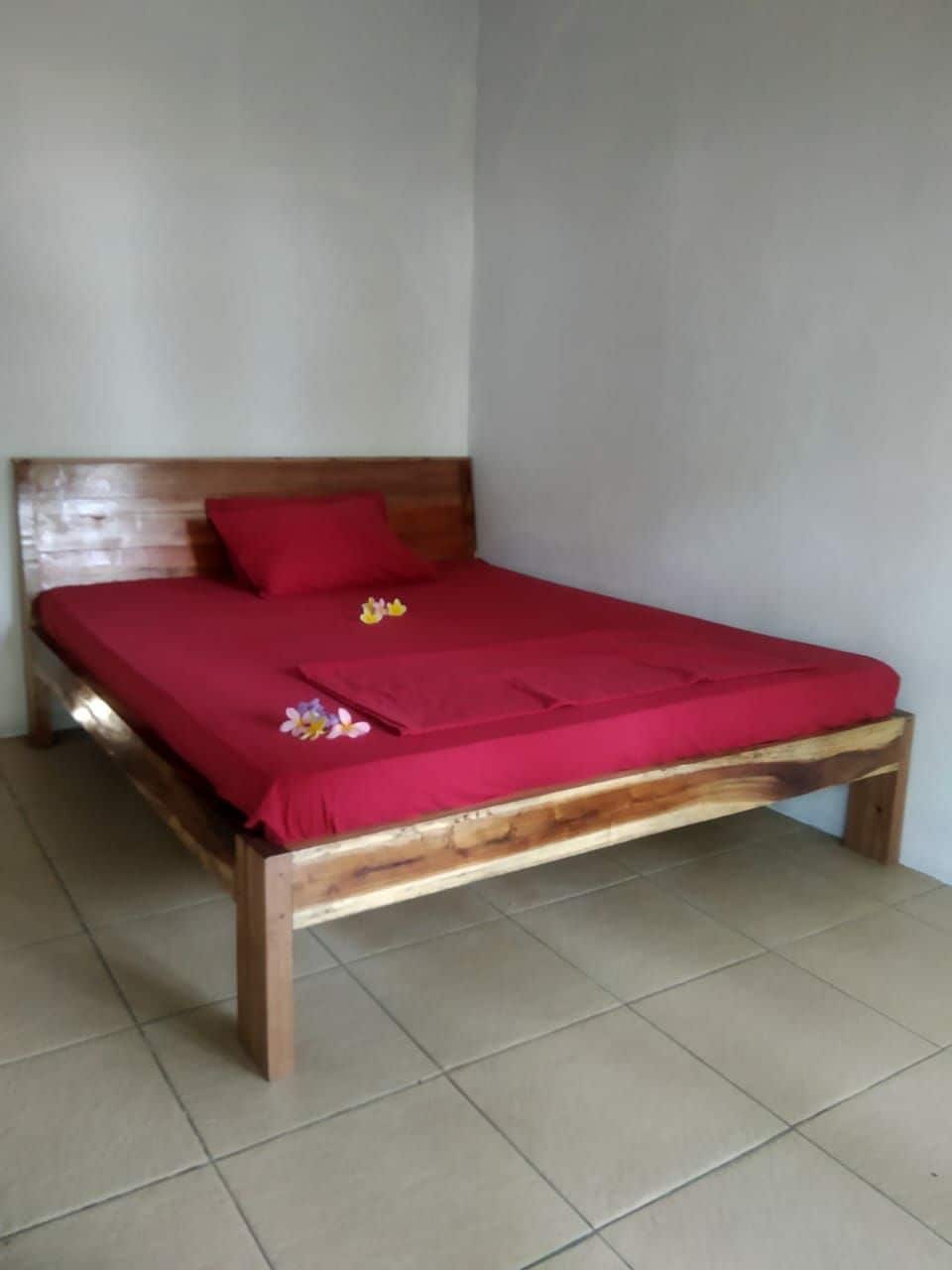 The room features a queen size bed with a sturdy wooden frame, dressed in a vibrant red sheet and matching pillow. A simple yet serene atmosphere is created by the light-colored walls and tiled floor, allowing for relaxation and comfort.
