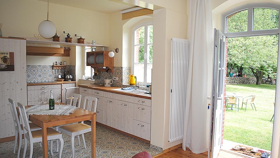 A bright kitchen area features white cabinetry, wooden countertops, and a dining table with several chairs. Large windows allow natural light to fill the space, while an open door provides a view of the garden outside, enhancing the connection to nature.