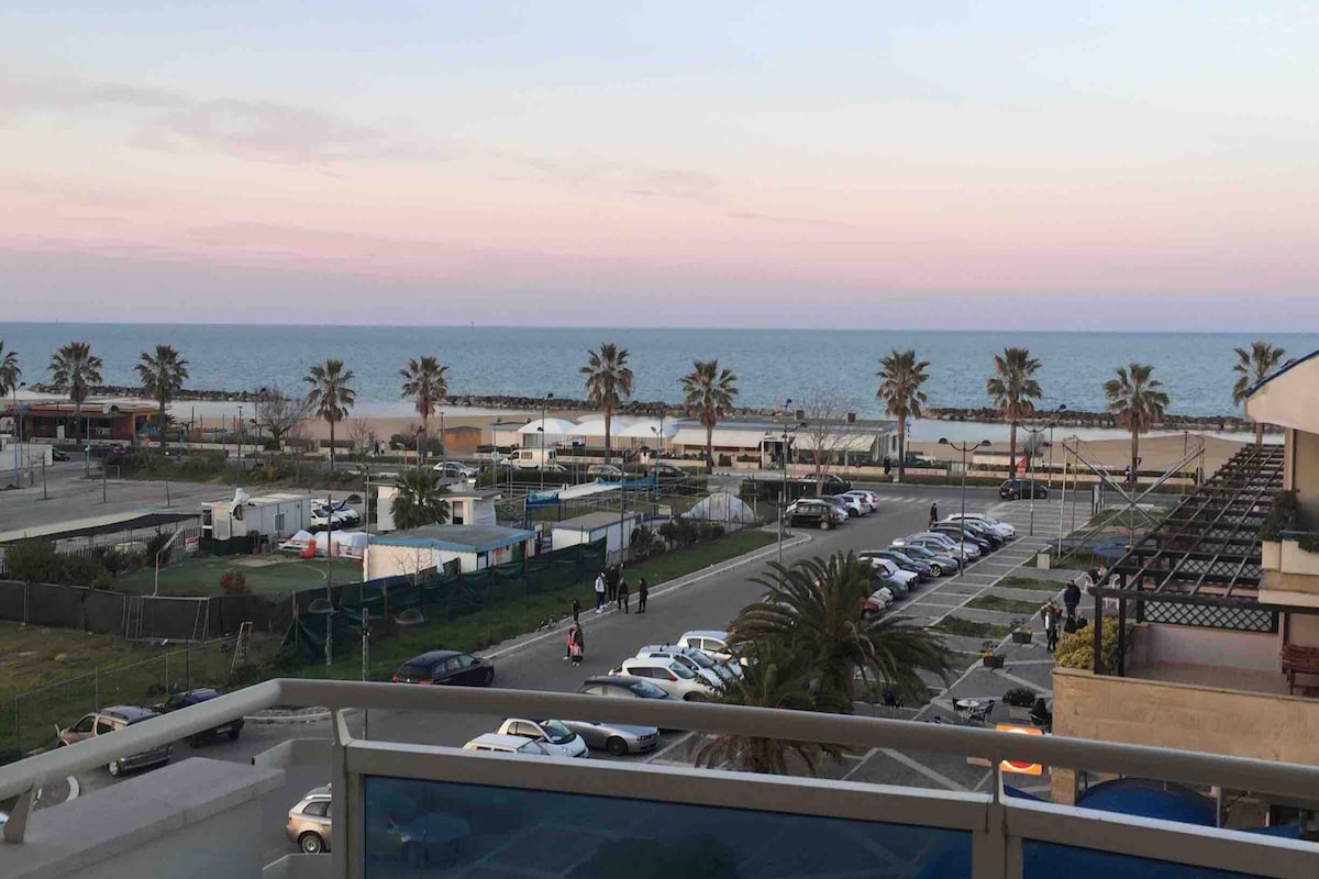 A wide view of the coastline is captured, showcasing sandy beach, palm trees, and the calm sea under a pastel sky. Vehicles are parked along the road, with some people visible strolling along the promenade, creating an atmosphere of leisure and community.