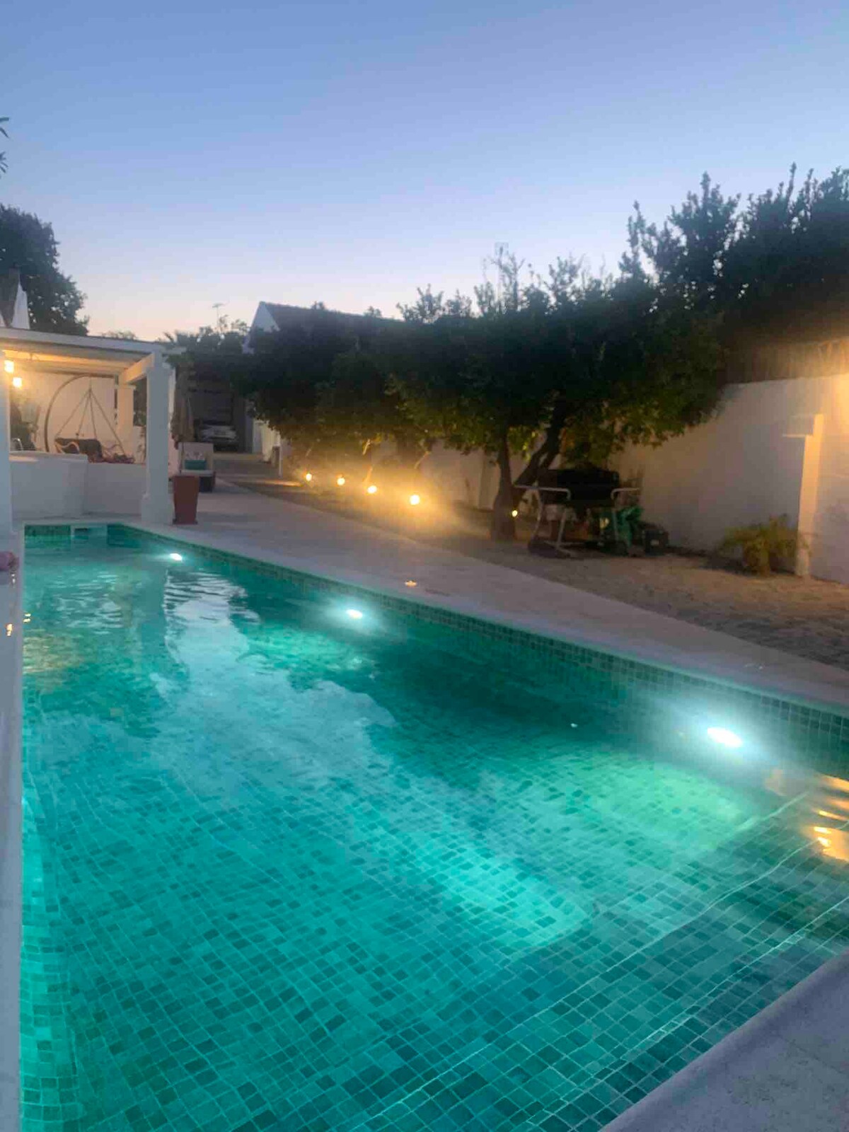 A tranquil swimming pool features illuminated turquoise tiles under the evening sky. Soft lighting lines the pool area, creating a serene ambiance. Surrounding fruit trees enhance the outdoor space, contributing to a peaceful environment suitable for relaxation.