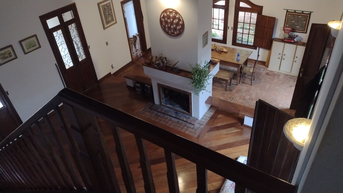A view from above captures the spacious living area, featuring a central fireplace surrounded by wooden accents. Light streams through large windows, illuminating the rustic furniture, which includes a small table and chairs. A decorative wall piece adds character to the warm atmosphere.