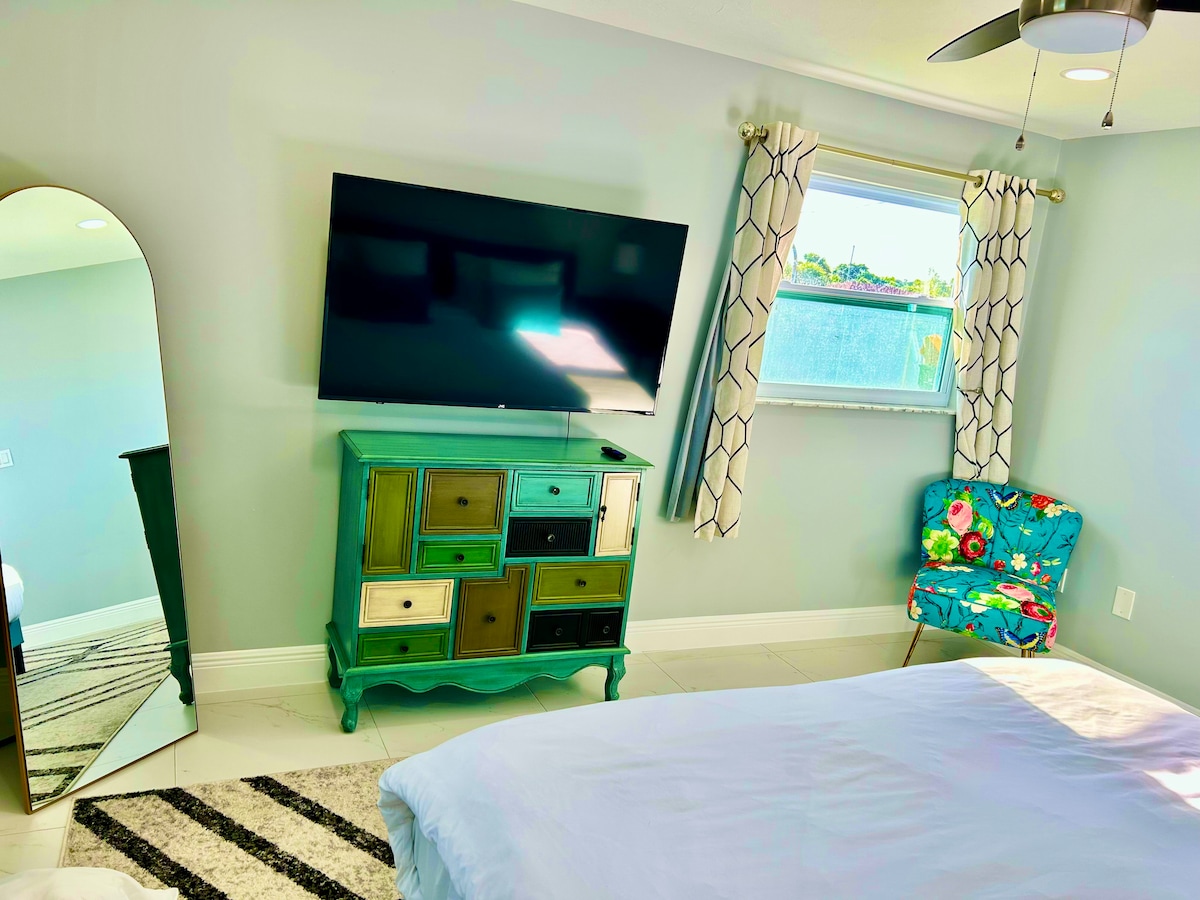 A light and airy bedroom features a king-size bed with crisp white bedding. A colorful accent chair adds vibrancy to the space. A mirrored dresser and a wall-mounted television enhance functionality. Natural light filters through the window, complemented by patterned curtains.