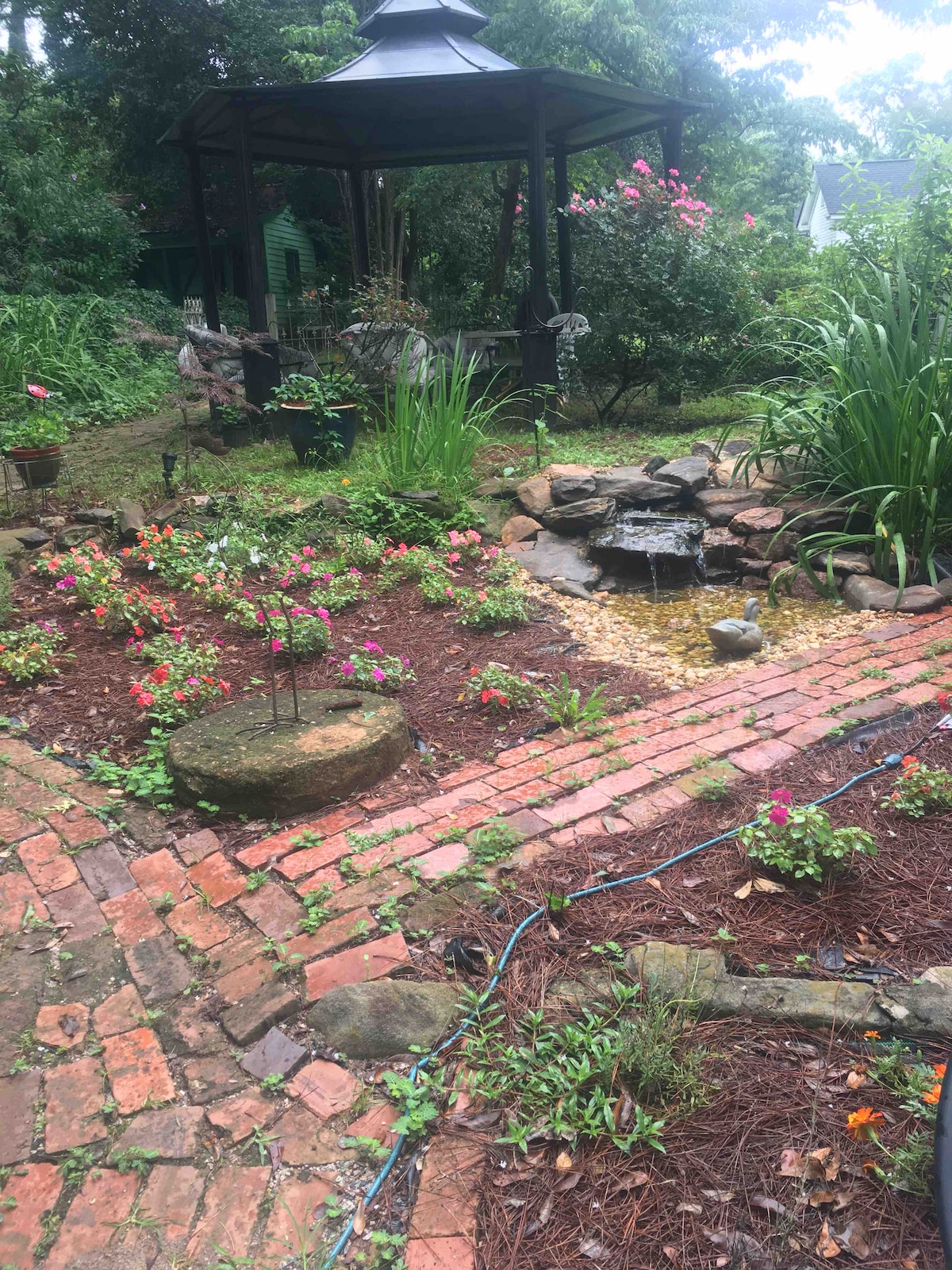 A serene garden setting features a stone path leading to a gazebo, surrounded by various flowering plants and greenery. A small pond with rocks is visible, adding to the tranquil atmosphere. The natural landscape is enhanced by soft lighting filtering through the trees.