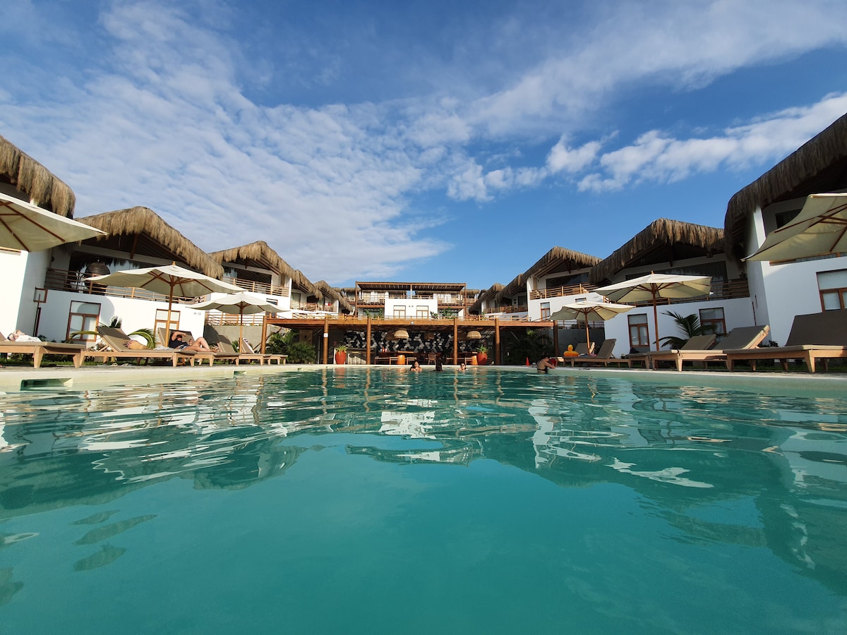 A clear view of a swimming pool is presented, reflecting the blue sky above. Sun loungers are arranged around the pool, complemented by umbrellas for shade. The background features a multi-level structure with a thatched roof, creating a welcoming atmosphere.