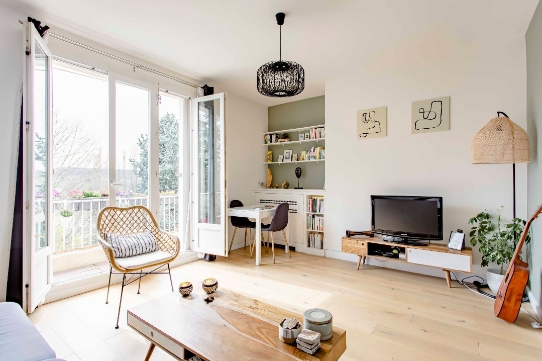 A bright living room is presented, featuring two large glass doors that open to a balcony, allowing natural light. A round, woven chair is positioned beside a wooden coffee table. A television sits on a low cabinet, and a dining table is visible against the wall.