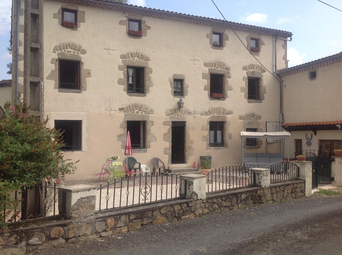 The exterior of the building features a two-story structure with traditional stone detailing. A small patio area is visible, furnished with a few chairs and a table. The surroundings include a gravel pathway, low fencing, and greenery, providing a serene rural atmosphere.