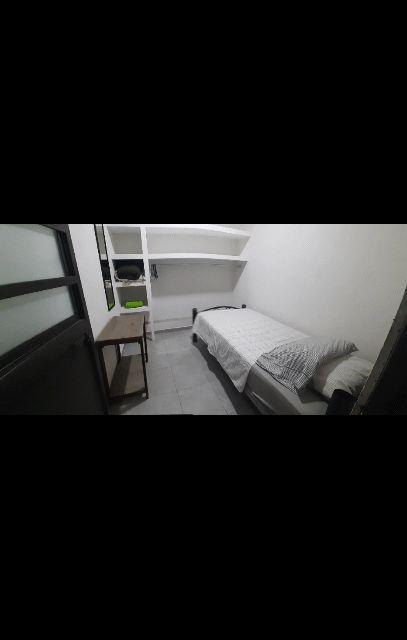 A compact room features a single bed with a light grey blanket, positioned against a white wall. A small shelving unit is mounted above the bed, providing storage space. Light-colored floor tiles and a nearby window contribute to an open feel.