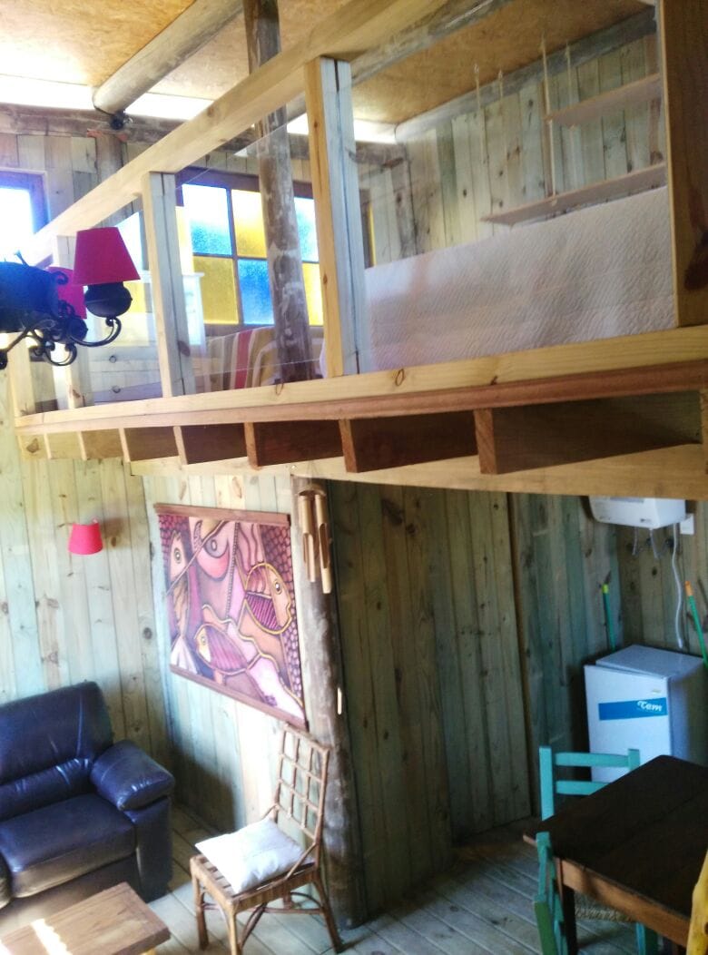 A lofted sleeping area is visible above in a wooden cabin, featuring a simple bed with a light-colored quilt. The ground level showcases a cozy seating area with a dark leather couch, a colorful wall hanging, and a small refrigerator. Natural wood walls enhance the rustic atmosphere.