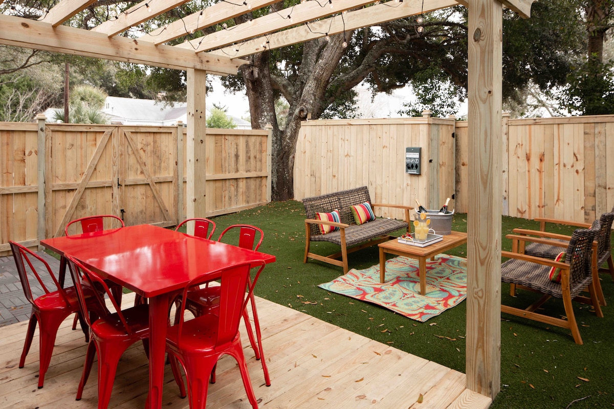 An outdoor seating area features a red dining table with matching chairs, surrounded by a green lawn. A cozy seating arrangement offers comfortable chairs and a patterned rug. Warm lights hang overhead, enhancing the relaxed ambiance in this private space.