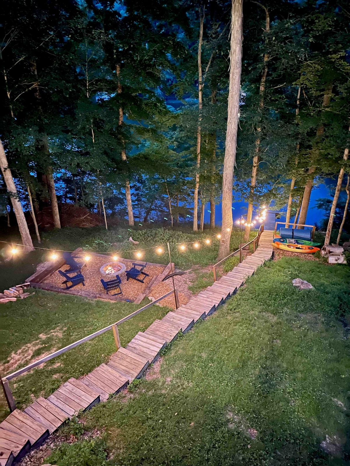 A wooden pathway bordered by lights leads to a gathering area with four chairs around a fire pit. Lush greenery surrounds the space, providing a serene atmosphere, while glimpses of the lake can be seen in the distance.