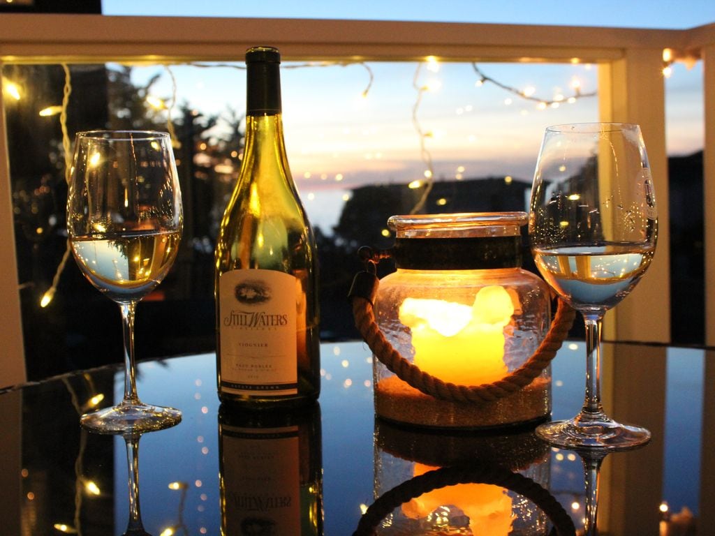 A bottle of white wine is displayed on a reflective surface, accompanied by two elegant wine glasses filled with wine. A softly glowing candle is positioned beside the wine, surrounded by twinkling string lights in the background, with ocean views visible as the sun sets.