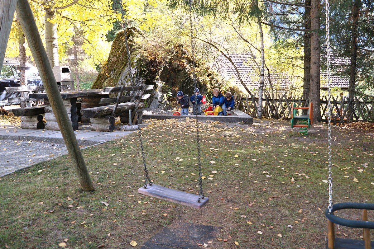 A tranquil garden area features a wooden swing hanging from a sturdy frame, surrounded by fallen leaves. In the background, a picnic table and benches are visible, while children interact with colorful play items, creating an inviting space for family activities.
