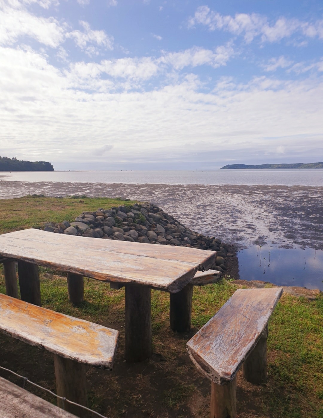 A rustic wooden table and benches are set on a grassy area, overlooking a tidal inlet. The calm water reflects the sky, which is adorned with clouds. Nearby, a rocky shoreline offers a natural texture against the greenery.
