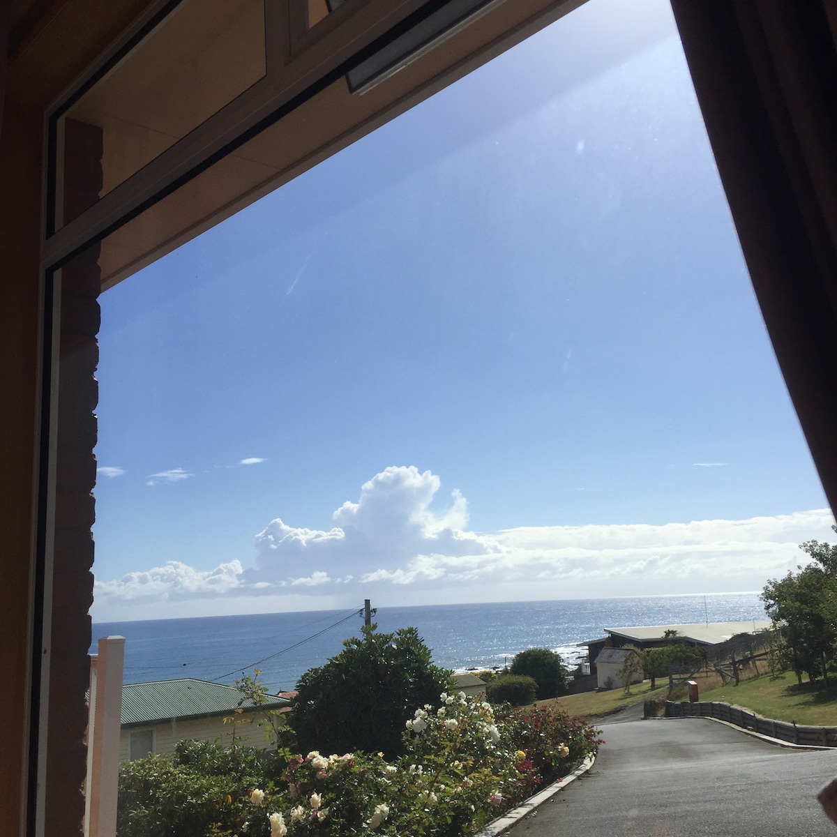 A view from a large window displays the ocean and sky, with gentle waves lapping at the shore. The scene features green hills and a few rooftops in the foreground, set under a bright blue sky dotted with clouds.