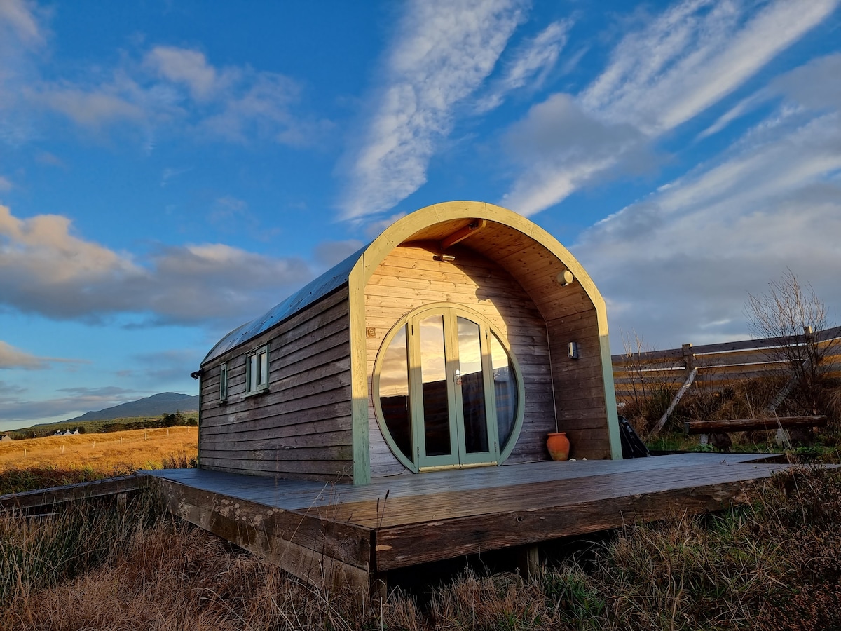 A Hobbit house featuring a distinctive round door is situated on a wooden deck amidst natural surroundings. The structure boasts a curved roof, and the exterior is crafted from wood. The sky above is filled with clouds, illuminated by the setting sun.