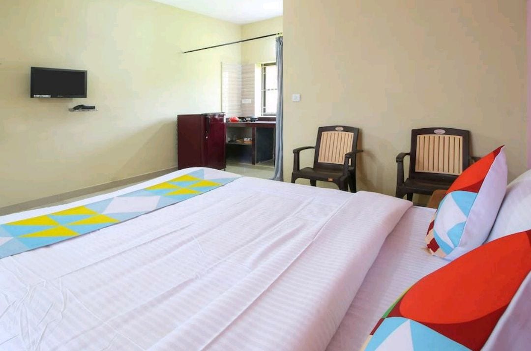 The spacious sleeping area features a neatly made bed with colorful accent pillows. Two wooden chairs are positioned nearby, offering additional seating. A television is mounted on the wall, and a wardrobe is visible in the background, creating a functional and inviting space.