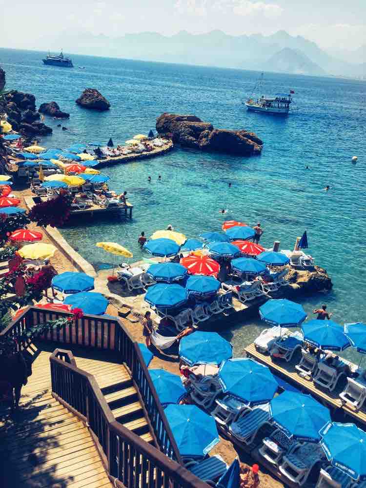 A scenic view of a beach area is captured, featuring numerous sun loungers arranged under vibrant blue and red umbrellas. The clear turquoise water reflects sunlight, and rocky formations are visible along the shoreline. Boats are anchored in the distance, against a backdrop of distant mountains.