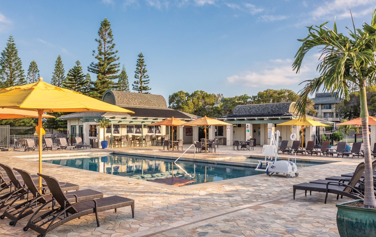 An outdoor swimming pool is surrounded by lounge chairs and large umbrellas, providing shaded seating areas. A poolside bar is visible in the background, along with a spacious deck that features a mix of palm trees and tropical greenery.