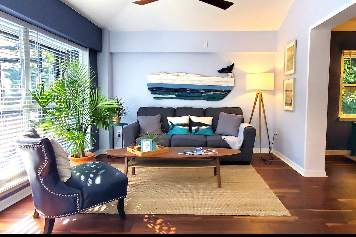 A bright living area showcases a mix of natural light and greenery. A comfortable gray sofa is paired with decorative pillows, while a wooden coffee table sits on a textured rug. A unique surfboard art piece adds character to the light blue walls.