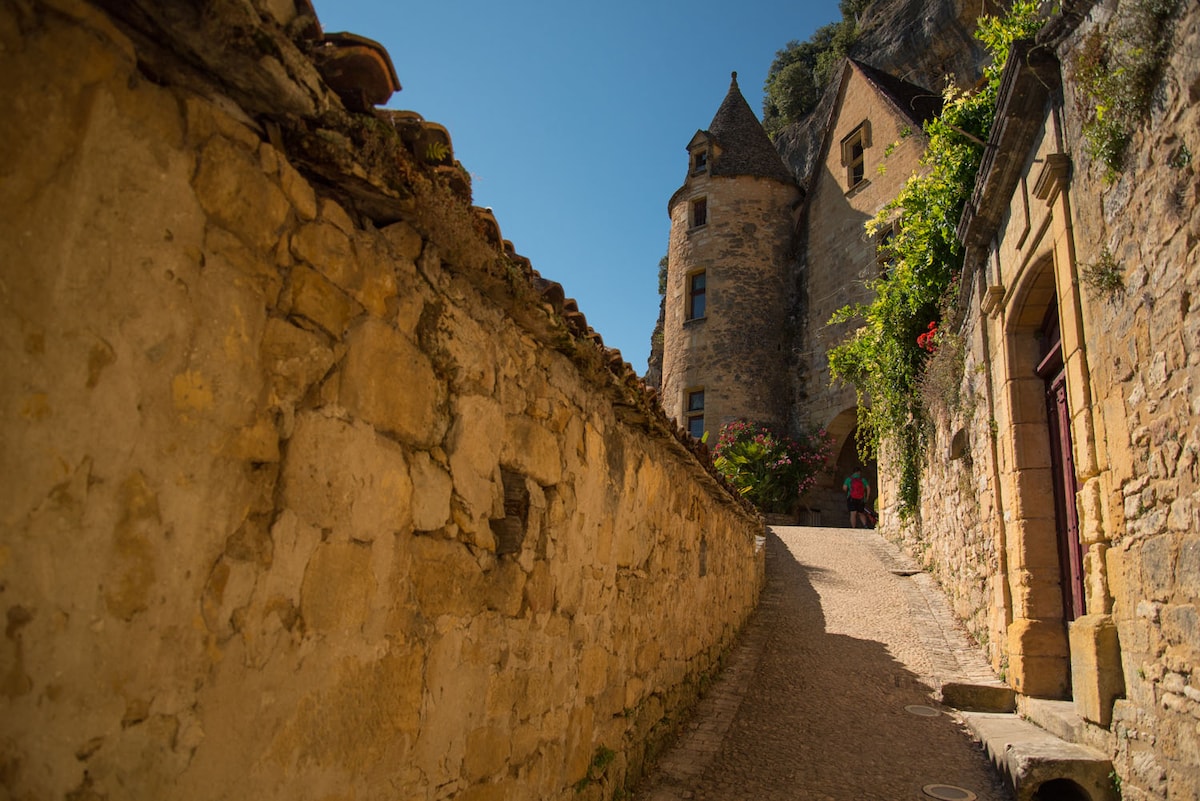 A winding path leads up a gently sloped street bordered by ancient stone walls. The surrounding buildings exhibit a blend of rustic charm, featuring turreted towers and vibrant greenery. Bright sunlight illuminates the scene, enhancing the texture of the weathered stone.