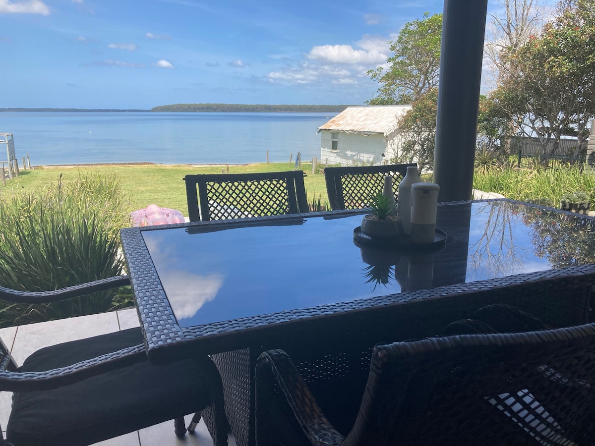 A dining table with four woven chairs overlooks a serene waterfront. The calm water reflects the clear blue sky and distant greenery. A small candle and decorative plant sit at the center of the table, enhancing the outdoor dining experience.