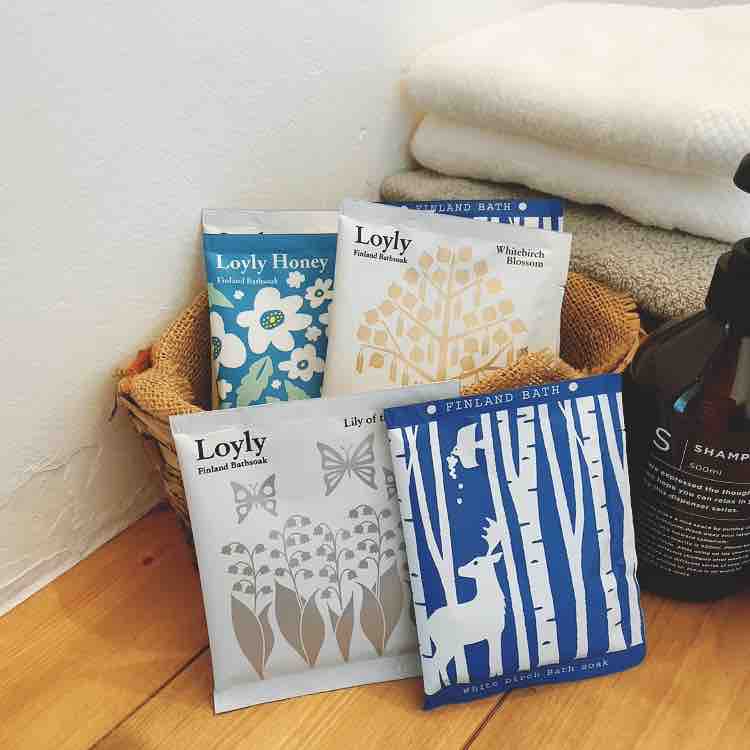 A collection of small, decorative packets of bath salts is displayed in a woven basket. The packets feature floral and nature-themed designs, each labeled with different bath names. Soft towels are arranged in the background, alongside a dark bottle of shampoo, enhancing the spa-like environment.