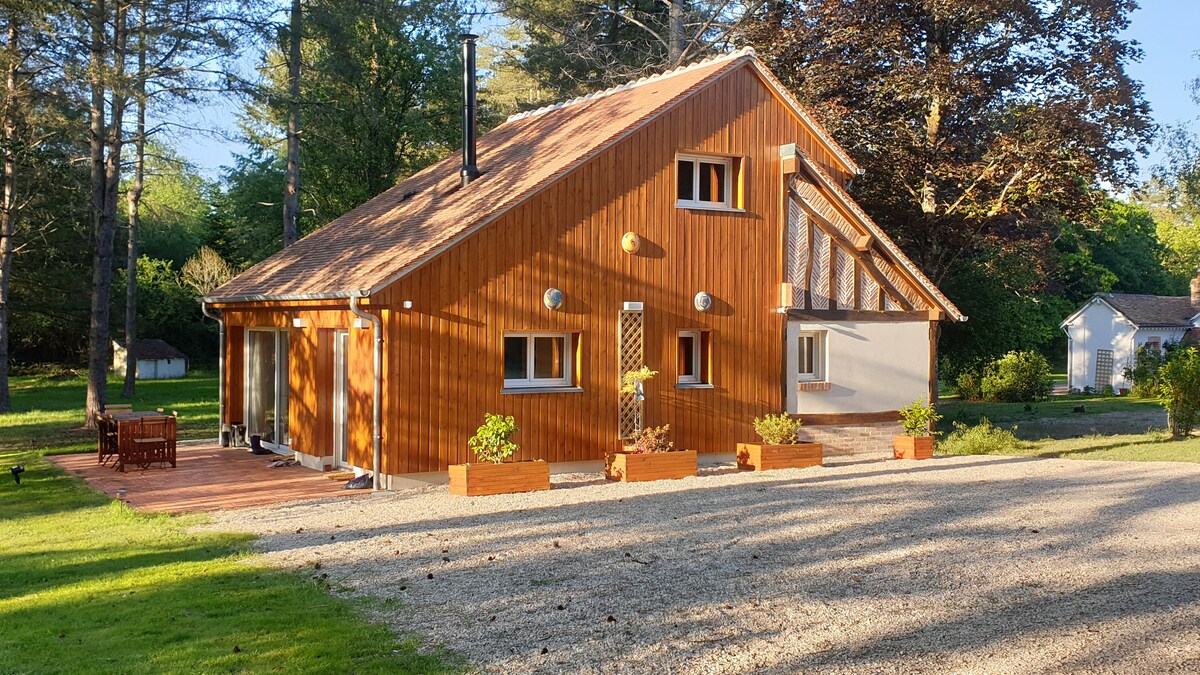 A wooden home is set amidst a spacious green landscape. The exterior features various windows allowing natural light, with decorative flower boxes lining the front. A gravel pathway leads to the entrance, bordered by well-kept grass and trees in the background.