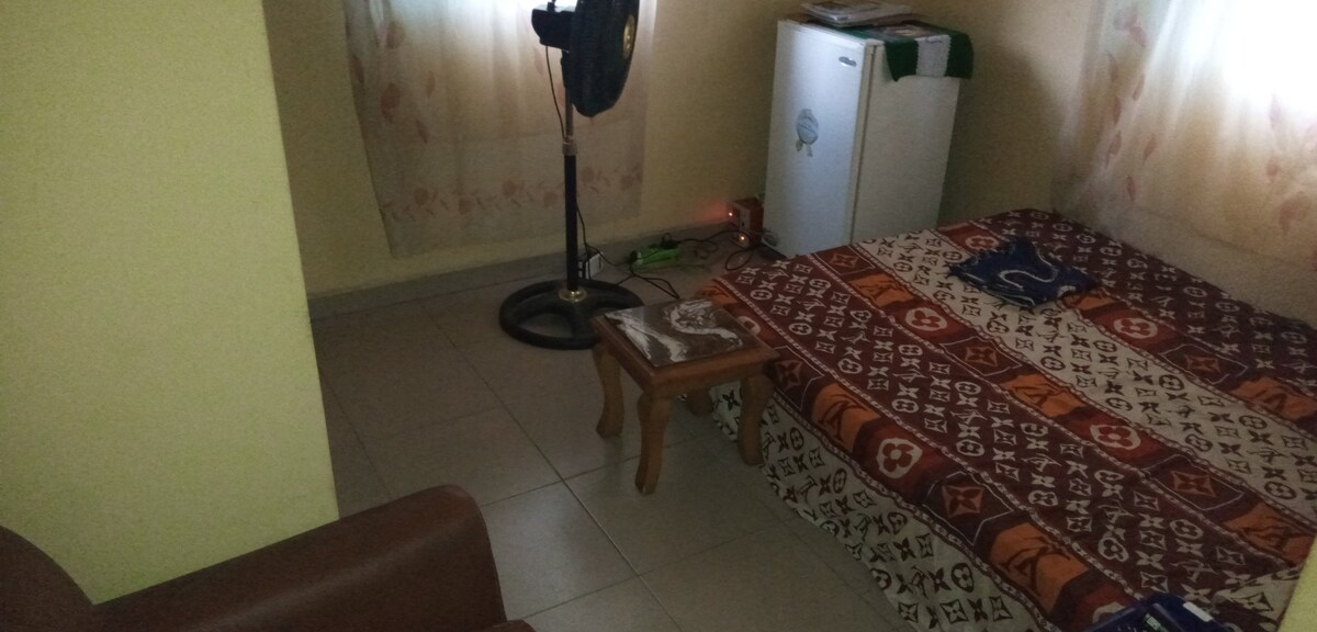 A cozy room is displayed, featuring a spacious bed adorned with patterned bedding. A small wooden stool is positioned nearby, alongside a fan and a refrigerator. Soft natural light filters through the sheer curtains, illuminating the tiled floor.