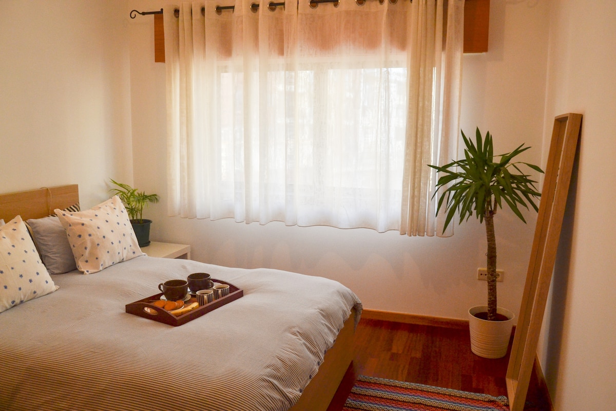 A bright bedroom is furnished with a spacious bed covered in light grey bedding. A wooden tray is placed on the bed, featuring various cups and small bowls. Natural light filters through the sheer curtains, illuminating a potted plant in the corner.