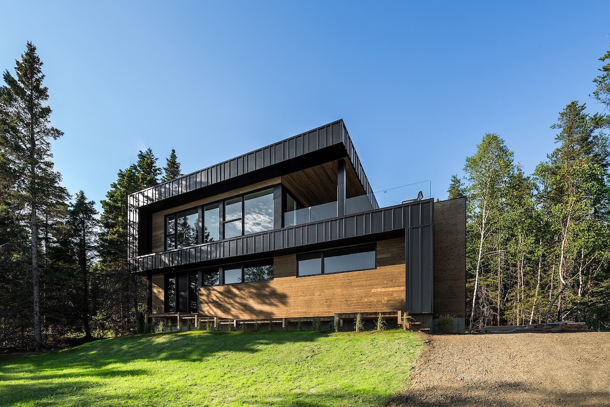 The modern villa is set against a natural backdrop, featuring a balanced combination of wood and modern materials. Expansive glass windows offer views of the surrounding greenery, while the upper terrace adds a serene outdoor space. The landscape includes well-maintained grass and trees.