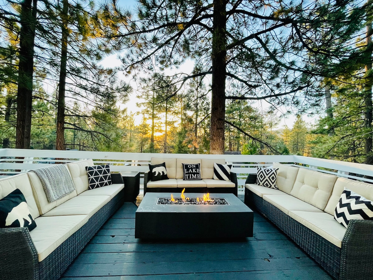 An outdoor seating area is surrounded by towering trees, featuring an arrangement of sectional sofas that invite relaxation. A modern fire pit sits at the center, providing warmth and ambiance. Decorative pillows add subtle accents to the design, complementing the natural surroundings.