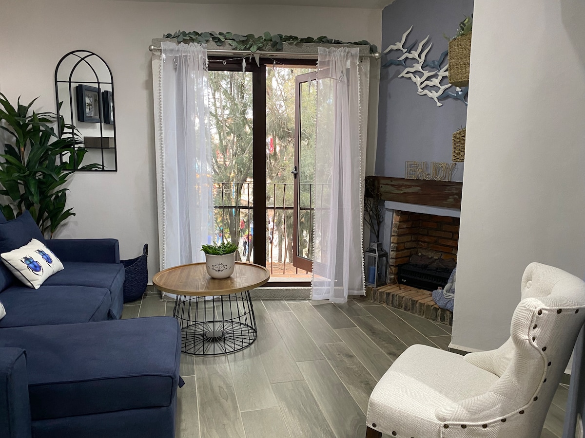 A bright living area features a blue sofa and a white accent chair. Large glass doors open to a balcony, allowing natural light to fill the space. A round wooden coffee table sits on a light wood floor, accompanied by a potted plant and decorative wall accents.