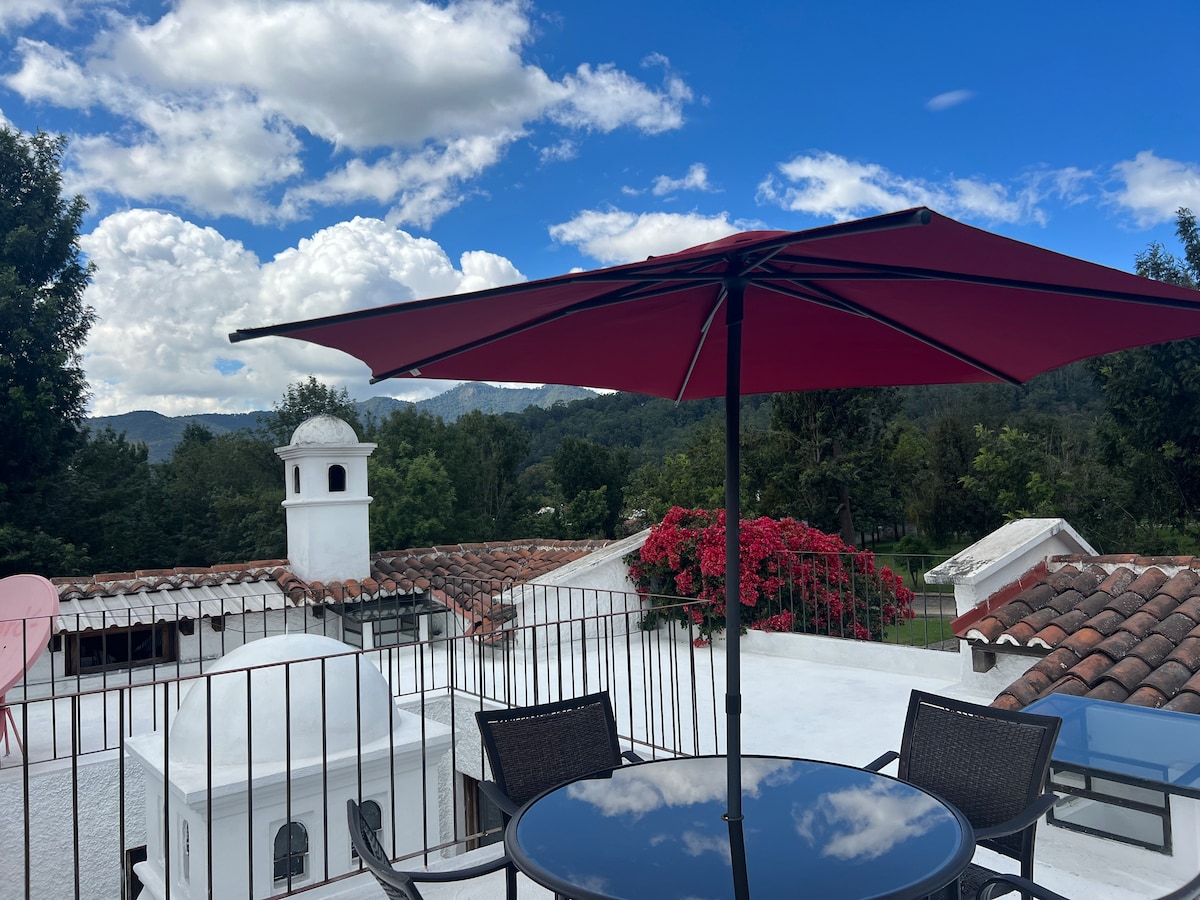 A rooftop terrace features a red umbrella shading a glass table surrounded by black chairs. Lush green hills and a bright blue sky with clouds create a serene backdrop. Red bougainvillea adds a touch of color along the railing, enhancing the peaceful outdoor setting.