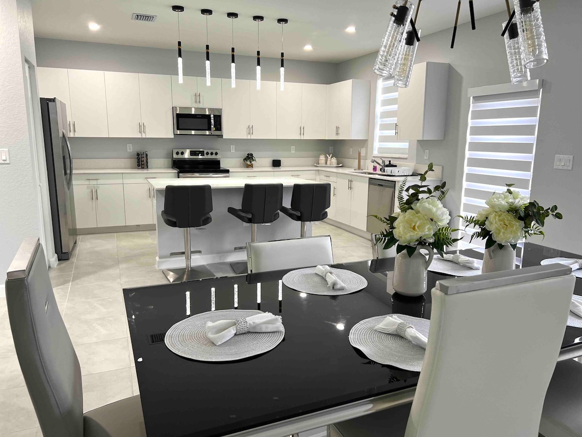 A modern kitchen and dining area feature sleek white cabinetry and stainless steel appliances. A glass dining table is complemented by comfortable chairs, set with elegant dinnerware. Pendant lights hang above the kitchen island, providing ample illumination for both cooking and dining.