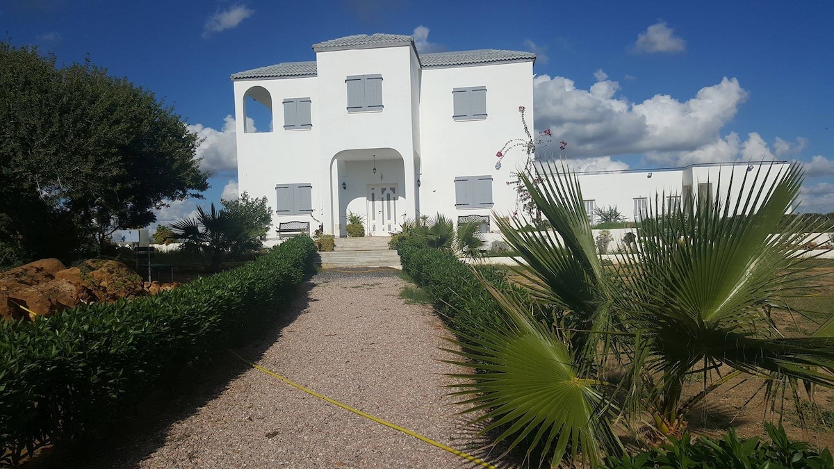 A charming white house is framed by lush greenery, with a gravel pathway leading to the entrance. Shuttered windows and a welcoming front door provide a glimpse of the property, set against a backdrop of blue sky and fluffy white clouds.