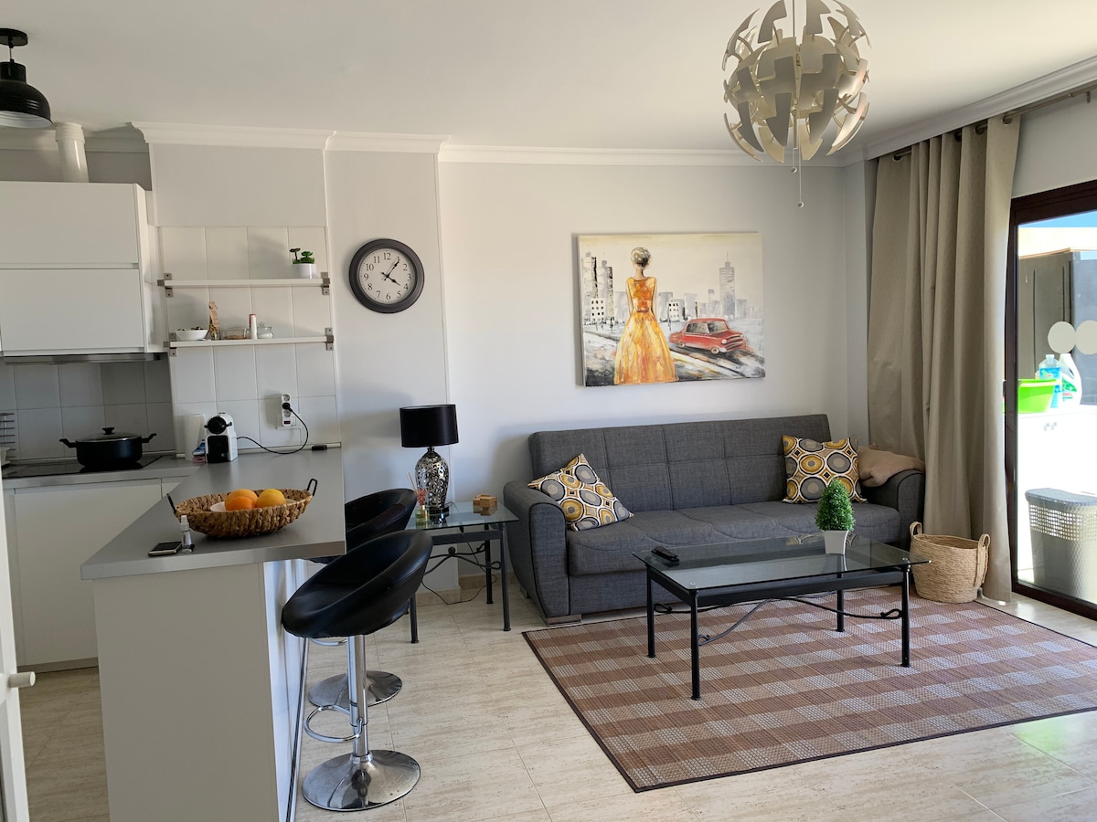 A modern living area features a comfortable gray sofa adorned with decorative cushions. A sleek glass coffee table sits on a patterned rug, and a round lamp provides soft lighting. A wall-mounted clock and a colorful artwork add character, with a door leading to an outdoor space.