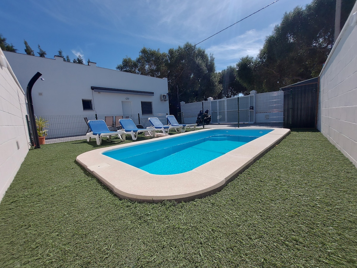 A private pool area is surrounded by a green lawn, featuring four lounge chairs positioned along one side. The pool is rectangular, with clear blue water reflecting the sky above, and a fence provides added privacy to the space.