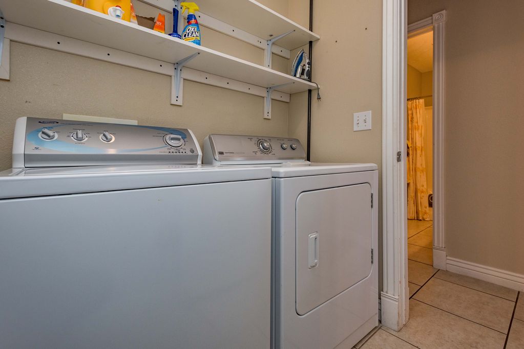 A laundry area is equipped with a washing machine and dryer, positioned next to elevated shelves holding laundry supplies. The tiled floor is visible, and a doorway in the background leads to another room, highlighted by soft lighting.