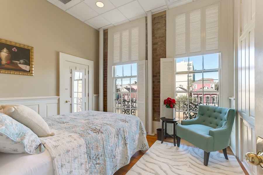 A bright bedroom features a spacious bed covered with a light-patterned quilt. Large windows allow natural light to fill the room, and a green accent chair is positioned beside a decorative area rug. A piece of artwork adorns the wall above the bed.