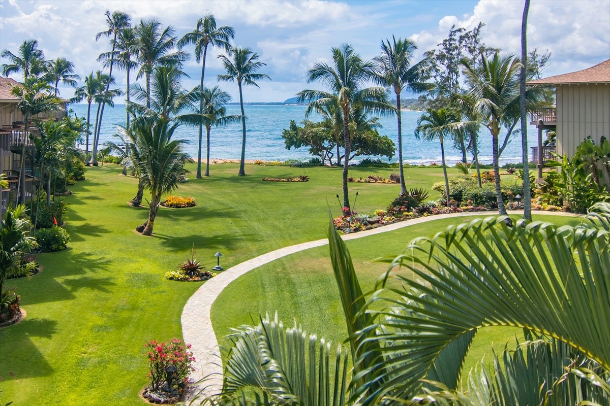 A lush, landscaped lawn is bordered by palm trees and tropical plants, leading to the ocean. The serene blue waters are visible in the background under a partly cloudy sky, providing a tranquil setting for relaxation.