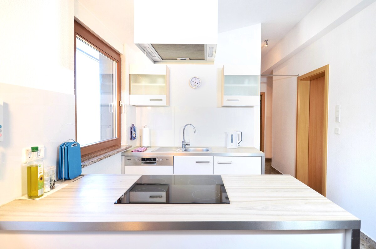 A modern kitchen is equipped with sleek cabinetry and a spacious countertop. A stove is integrated into the island, with a sink and various appliances visible. Natural light filters through the window, illuminating the clean, organized space.