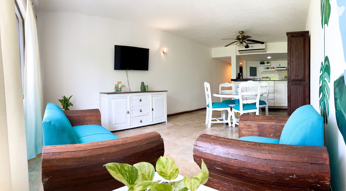 An open living area features two comfortable chairs and a white cabinet. A flat-screen TV is mounted on the wall, opposite a dining table with white chairs. The space is illuminated by natural light from large windows, creating a bright and airy atmosphere.