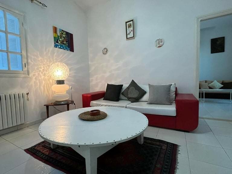 A bright living space features a white coffee table at the center, surrounded by a red and white sofa adorned with cushions in neutral tones. A unique lamp casts soft patterns on the wall, while two framed artworks add subtle color. Natural light filters through a window.