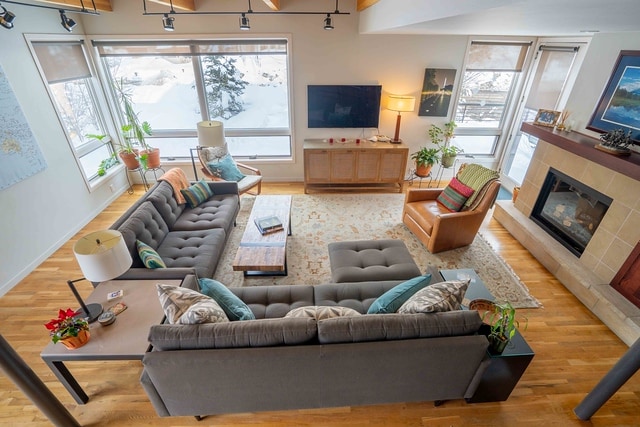 A spacious living area is illuminated by natural light from large windows. Comfortable sofas are arranged around a central coffee table, with plants and decorative accents visible. A gas fireplace stands on one side, and a television is mounted on the opposite wall, adding to the inviting ambiance.