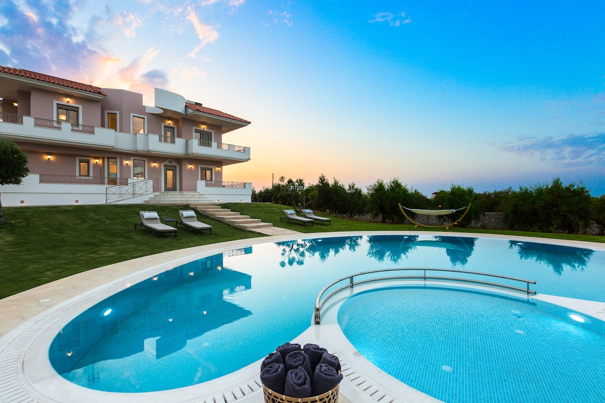 The image captures a luxurious villa set against a vibrant sky at twilight. A pristine oval pool reflects the sunset hues, surrounded by neatly arranged sun loungers. Lush greenery frames the scene, while a hammock adds a relaxing element to the outdoor space.