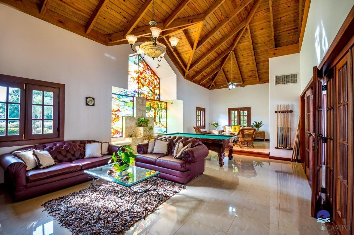 An open living space features a high wooden ceiling with exposed beams, while large windows allow natural light to illuminate the area. A plush leather sofa and elegant armchairs surround a coffee table atop a soft rug, and a billiard table is positioned nearby.